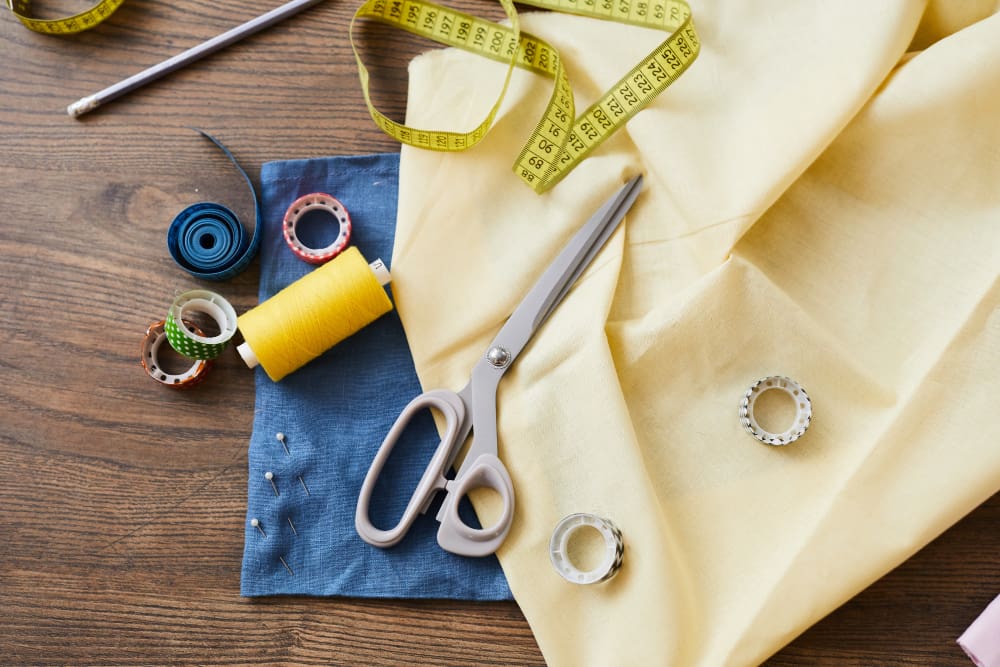 A pair of scissors, yellow fabric, measuring tape, thread spools, sewing pins, and a blue cloth arranged on a wooden table.