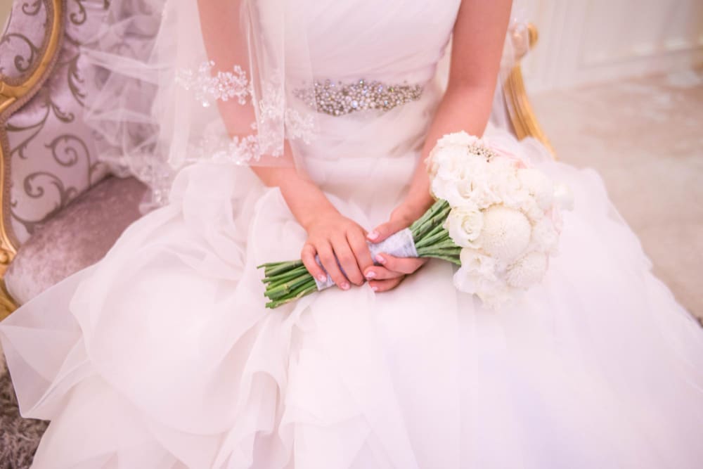 A bride in a white wedding dress sits holding a bouquet of white flowers in her lap.