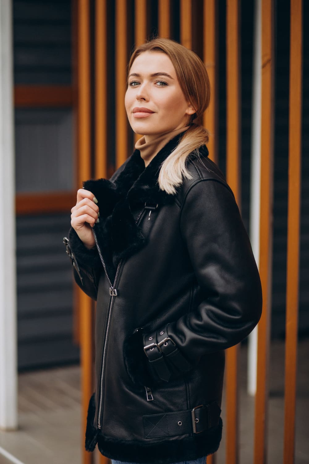 Woman standing outdoors, wearing a black leather jacket with fur lining, looking at the camera. Vertical wooden slats and grey wall are in the background.