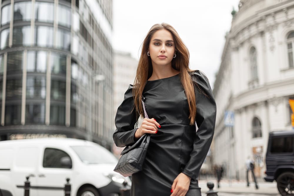 A woman in a black leather dress stands outdoors in a city street, holding a black handbag, with buildings and vehicles in the background.