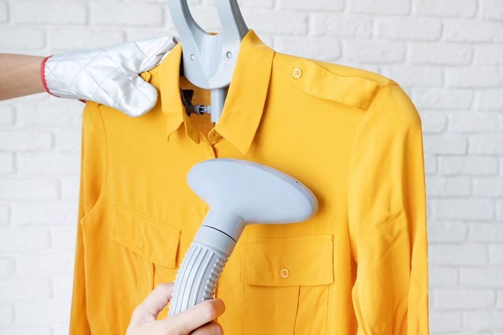 A person in an oven mitt steams a yellow button-up shirt on a hanger in front of a white brick wall.