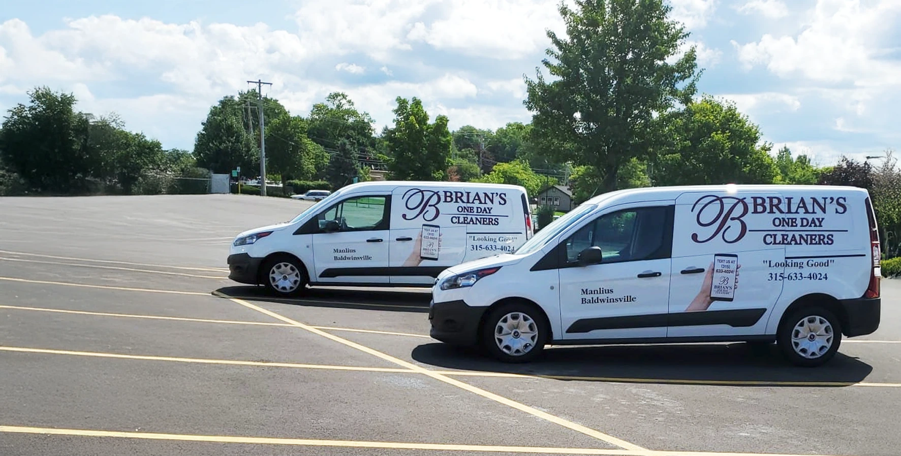 Two white vans with "Brian’s One Day Cleaners" branding are parked in an empty lot on a sunny day, with trees and clouds in the background.