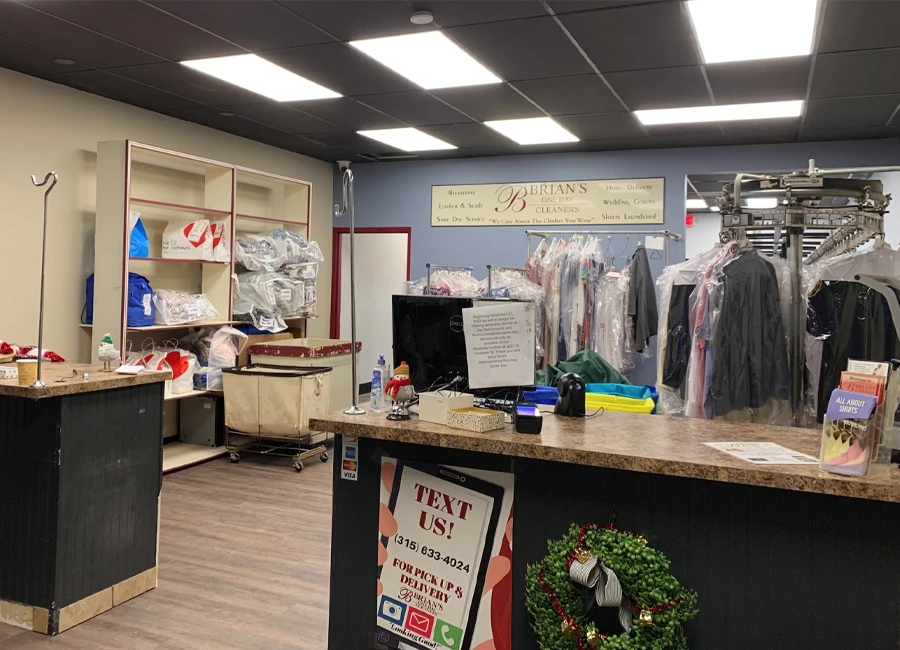 Interior of a dry cleaning store with counters, laundry racks holding clothes, shelves with supplies, and a sign displaying services behind the counter.