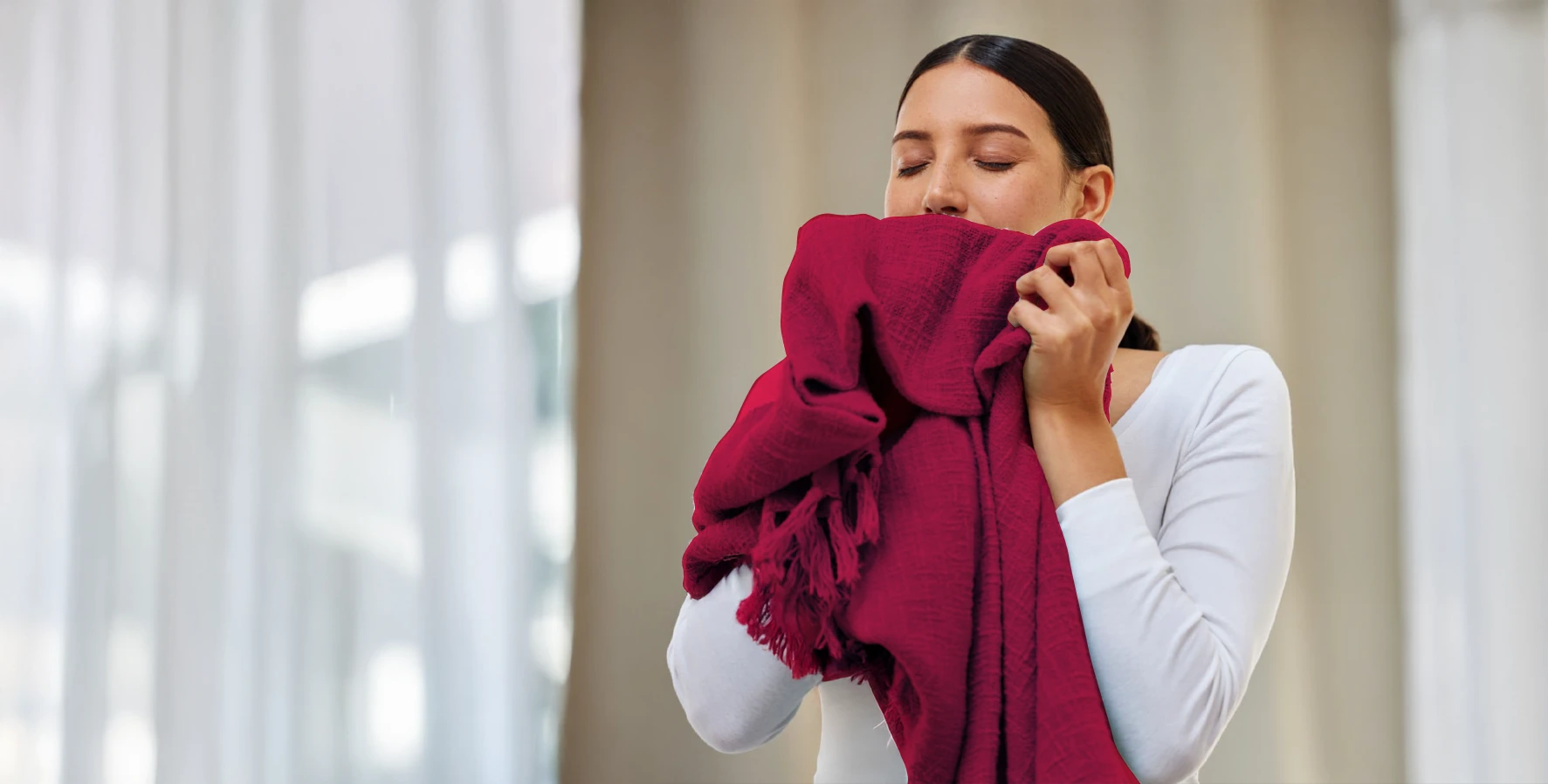 A woman in a white top holds and smells a folded red blanket or scarf, standing indoors with blurred curtains in the background.