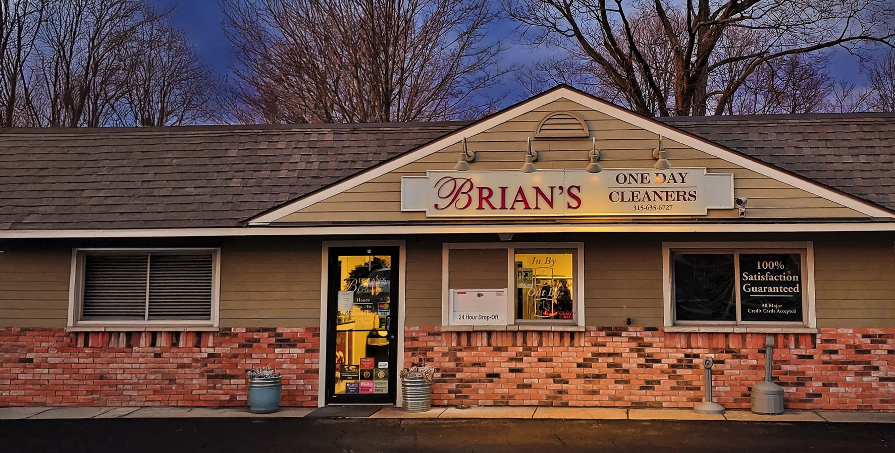 Exterior of Brian's One Day Cleaners with a lit entrance door, signage, and large display window at dusk.