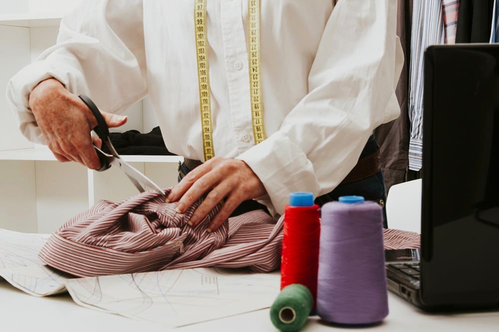 A tailor cuts striped fabric with scissors over a pattern, with thread spools and a laptop on the table; a measuring tape hangs around their neck.