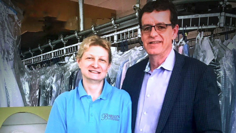 Two people stand in front of a conveyer with hanging dry-cleaned clothes; one wears a blue polo shirt with a “Bryan’s Cleaners” logo, the other wears glasses and a suit jacket.