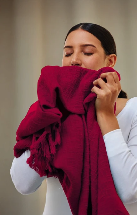 A woman in a white top holds and smells a red scarf, with her eyes closed and a neutral background behind her.