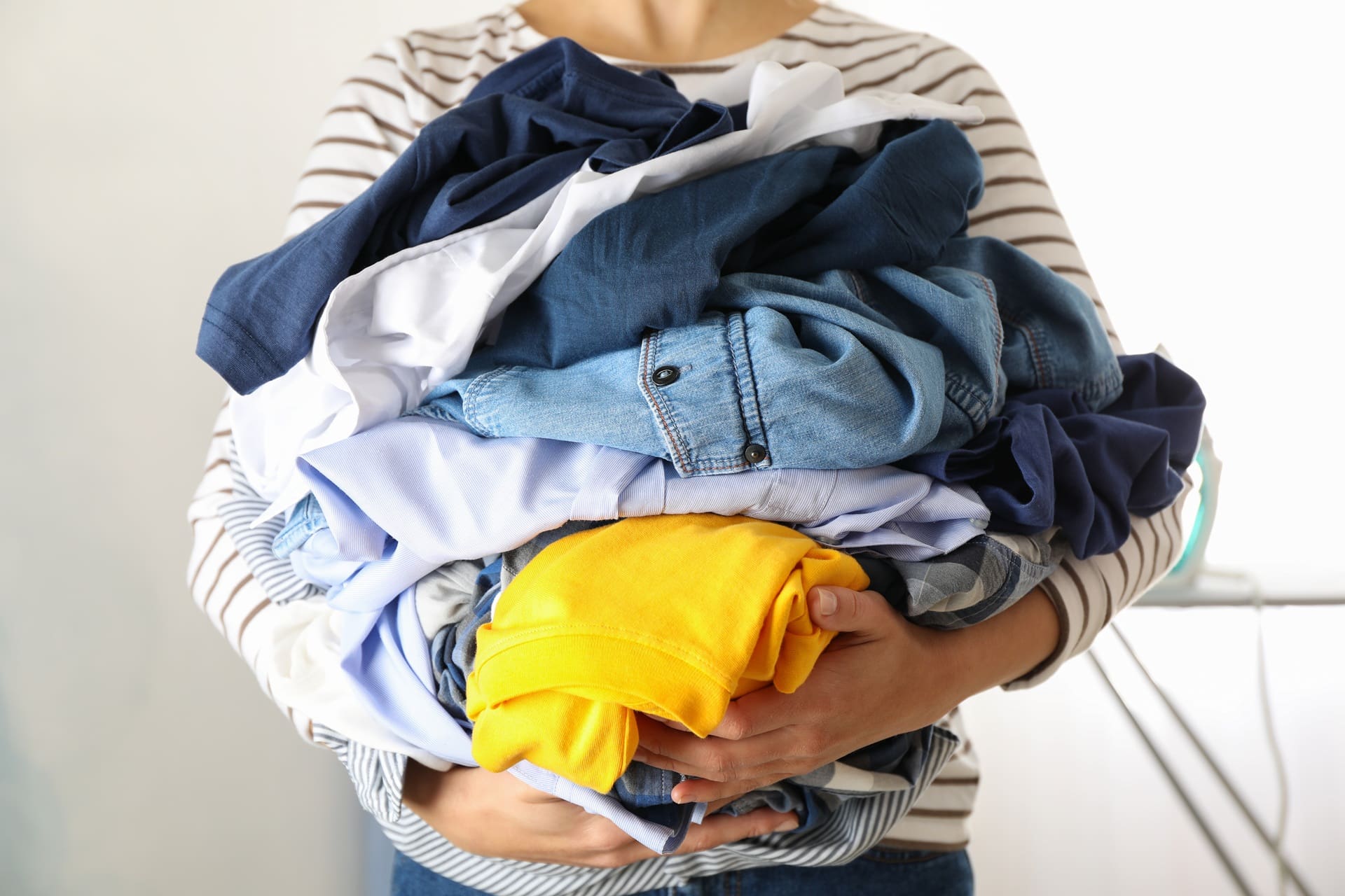 Person in a striped shirt holding a large pile of assorted clothes, including blue, white, and yellow garments.