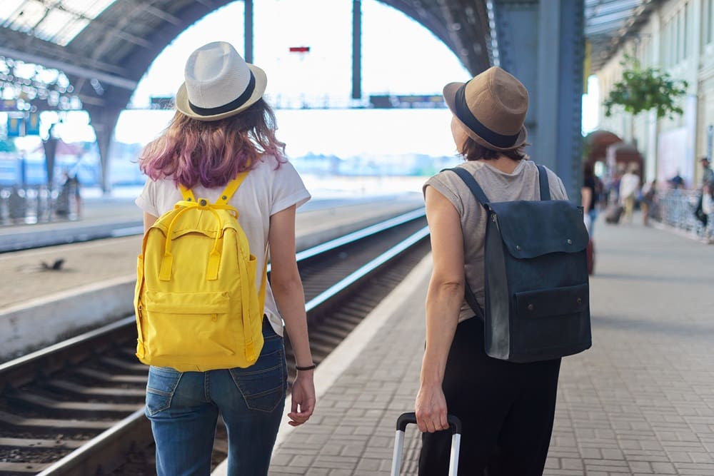 Two people with hats and backpacks stand on a train platform, one holding a suitcase, facing the tracks inside a large, covered railway station.
