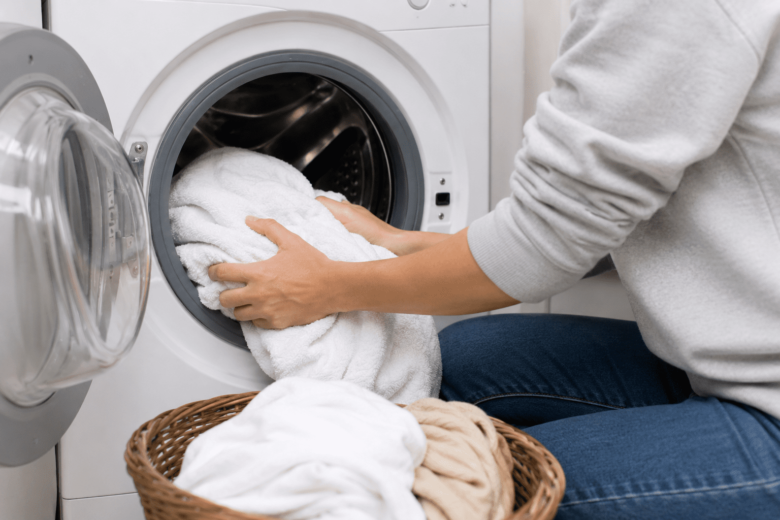 A person loads white towels into a front-loading washing machine, with a wicker laundry basket filled with clothes nearby.