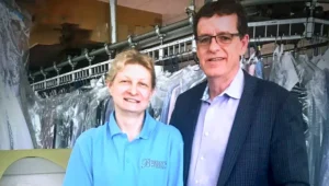Two people stand in a dry cleaning shop, with one wearing a blue polo shirt and the other in a suit. Garments in plastic covers hang on racks in the background.