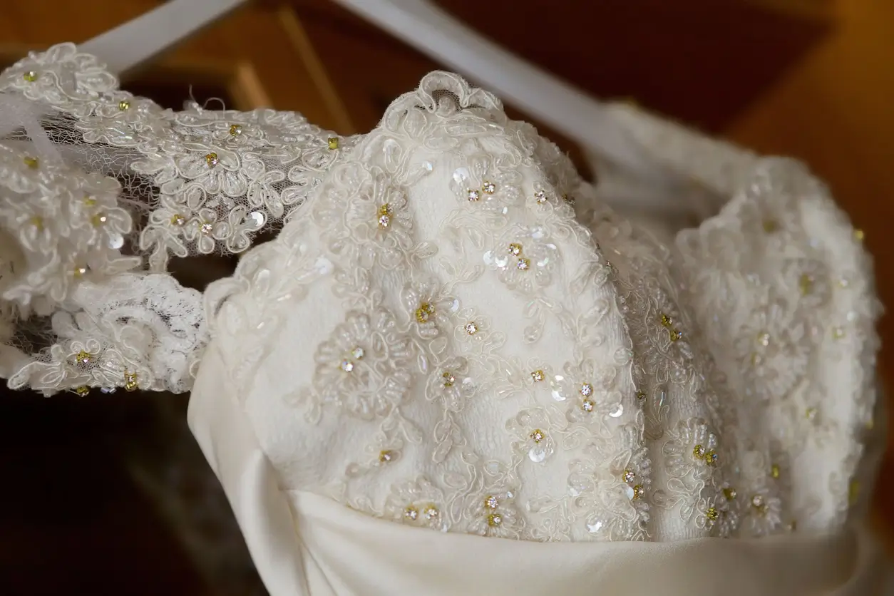 Close-up of an ornate white wedding dress hanging on a hanger, featuring lace, embroidery, and small bead details.